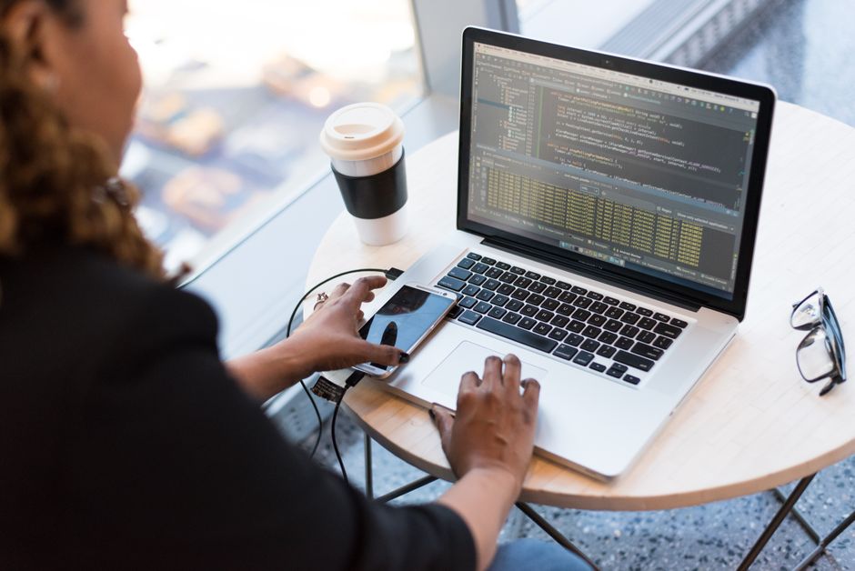 Femme noire programmant sur un ordinateur portable avec un café, un smartphone et des lunettes sur un bureau dans un environnement professionnel.