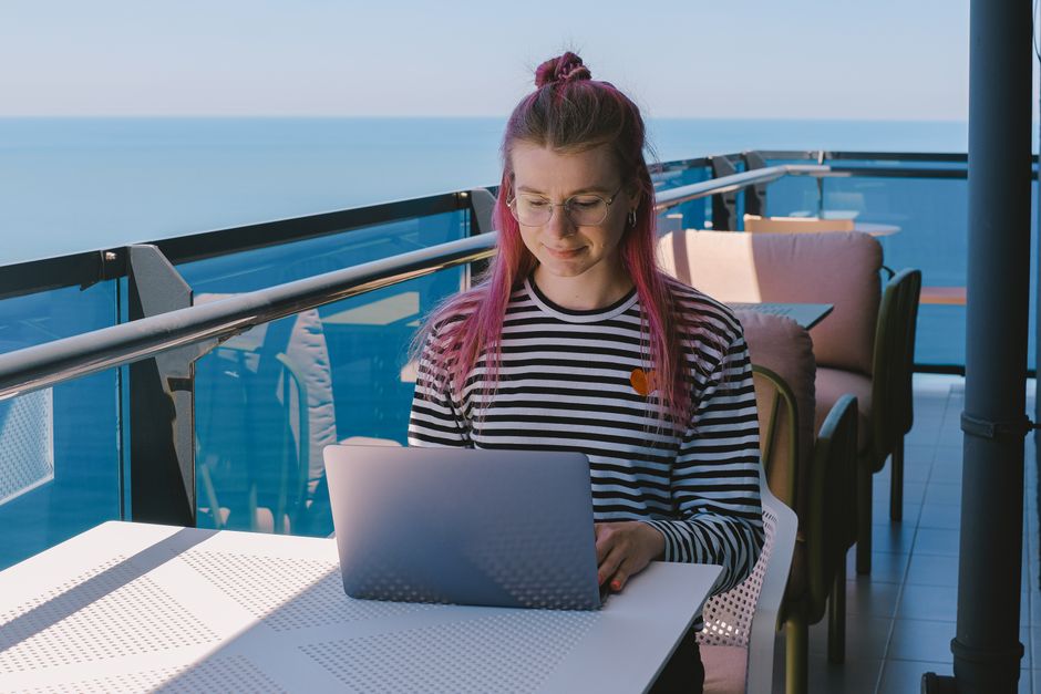 Jeune femme en télétravail sur un balcon ensoleillé face à l'océan, profitant d'un mode de vie nomade.