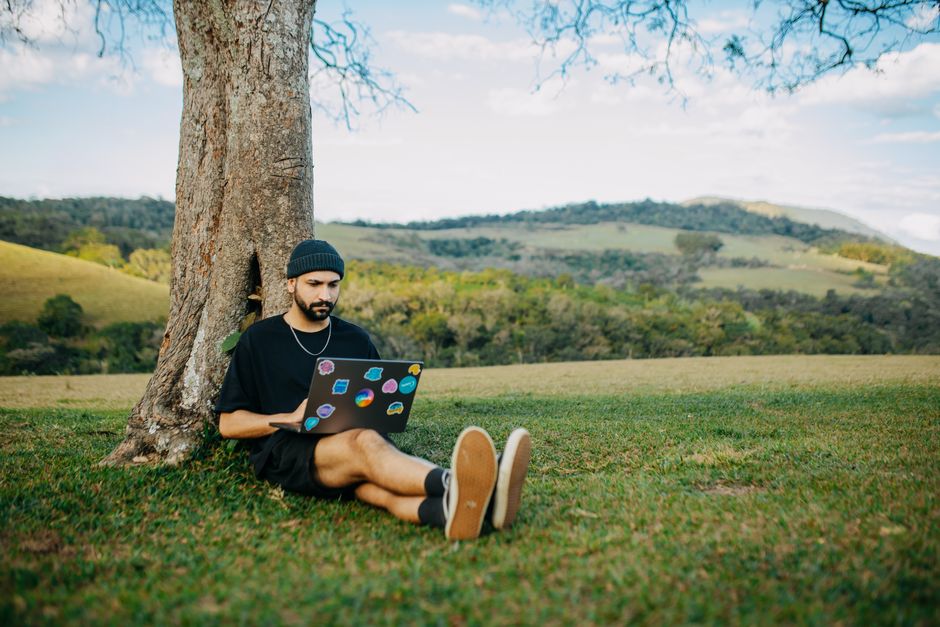 Jeune homme assis sous un arbre, travaillant sur un ordinateur portable dans une prairie paisible.
