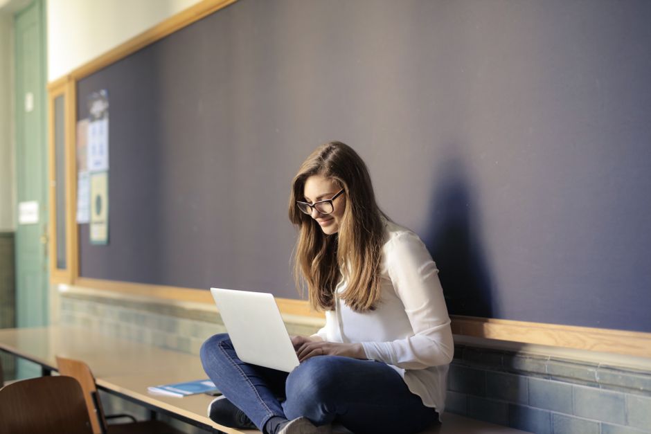 Une femme assise sur une table dans une salle de classe travaille sur son ordinateur portable, illustrant l'éducation moderne.