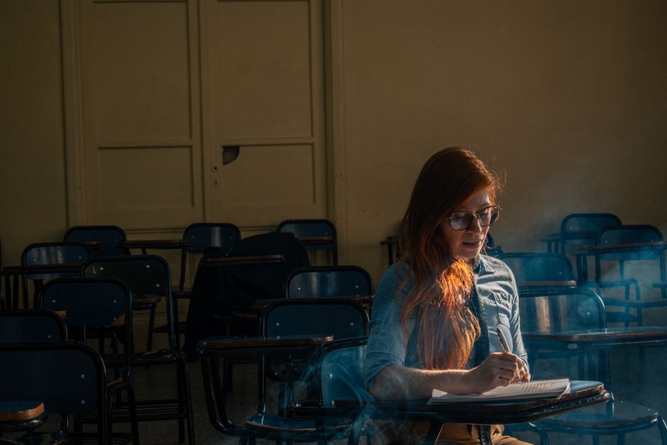 Une femme écrit dans un carnet dans une salle de classe vide, baignée par la lumière du soleil.