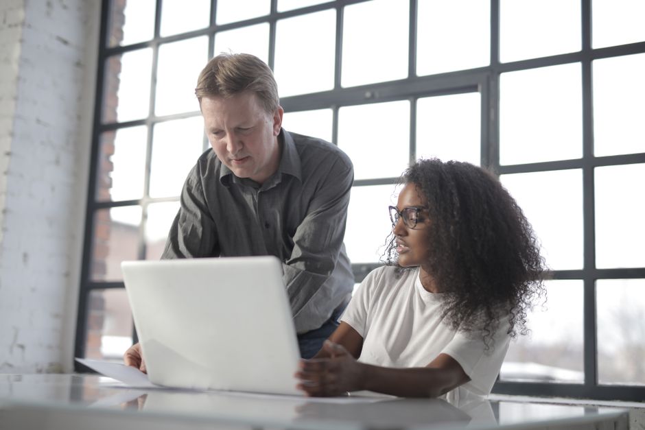 Entrepreneur professionnel consultant un ordinateur portable avec une collègue afro-américaine, étudiant des documents dans un bureau au style industriel devant une grande baie vitrée.