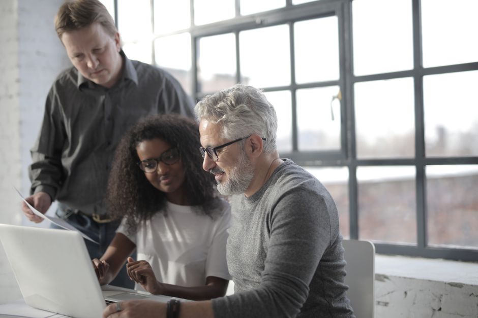 Équipe de professionnels travaillant ensemble avec un ordinateur portable et des documents dans un bureau au style industriel devant une grande fenêtre.
