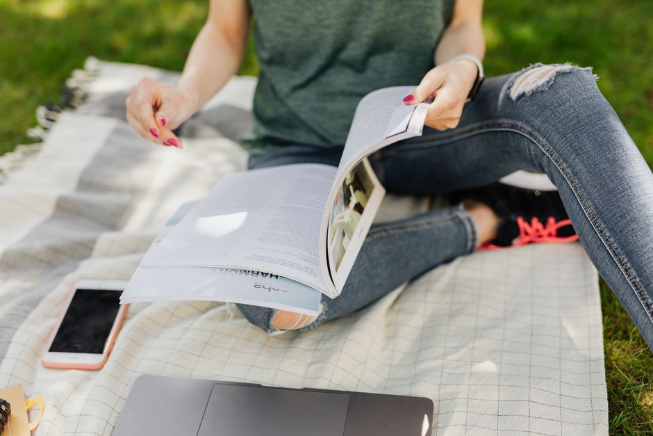 Gros plan sur une étudiante dont le visage n'est pas visible, feuilletant un manuel scolaire assise sur une couverture dans un parc verdoyant et calme en été, avec un téléphone portable et un ordinateur portable à ses côtés.