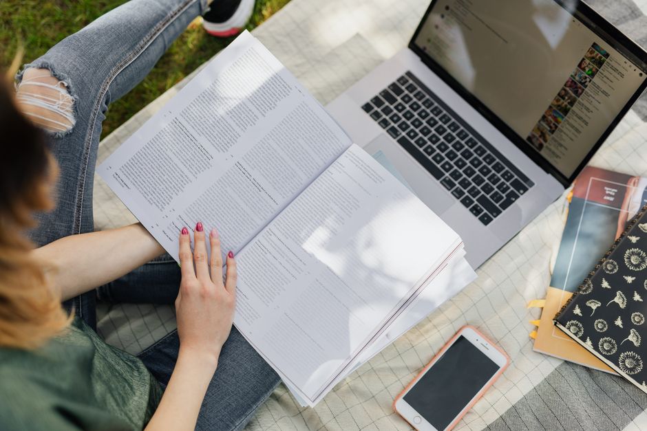 Vue en plongée d'une étudiante anonyme étudiant un manuel, assise avec un ordinateur portable et un smartphone dans un parc ensoleillé.