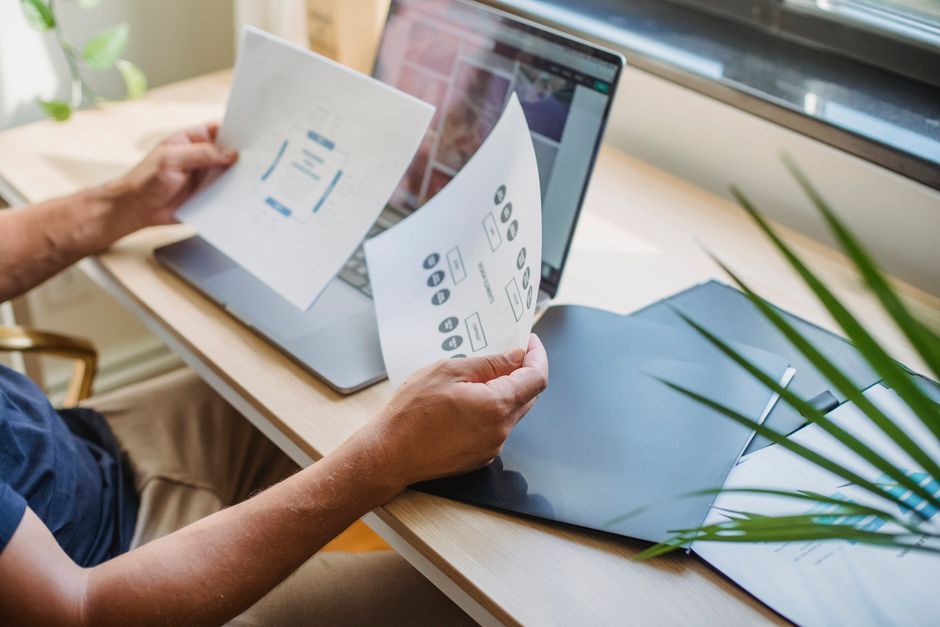 Designer masculin anonyme en tenue décontractée, assis à une table avec un ordinateur portable ouvert et des dossiers, lisant des documents.
