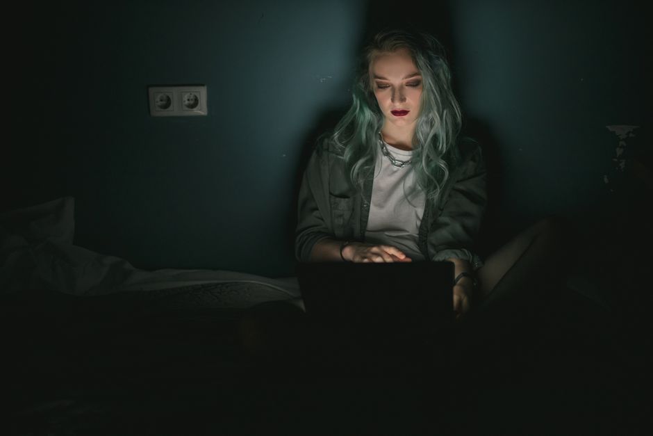 Femme aux cheveux colorés utilisant un ordinateur portable dans une pièce faiblement éclairée, travaillant tard le soir.