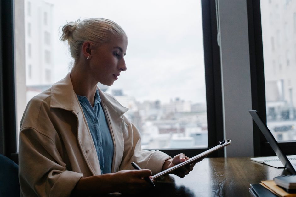 Vue de profil d'une jeune femme concentrée en blouse blanche, assise à un bureau en bois et consultant des rapports sur un presse-papiers.