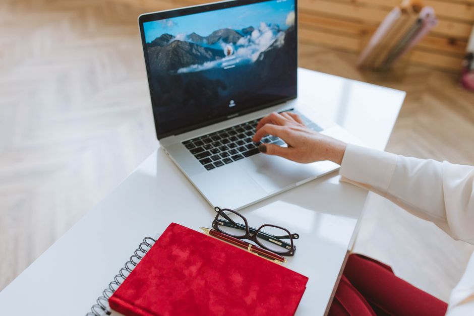 Personne travaillant sur un ordinateur portable avec un carnet rouge et des lunettes sur une table blanche.