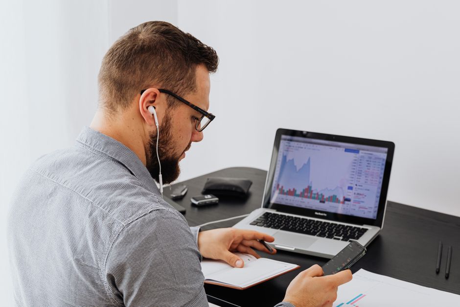 Homme d'affaires avec des écouteurs analysant des graphiques boursiers sur un ordinateur portable tout en utilisant un smartphone et un carnet.