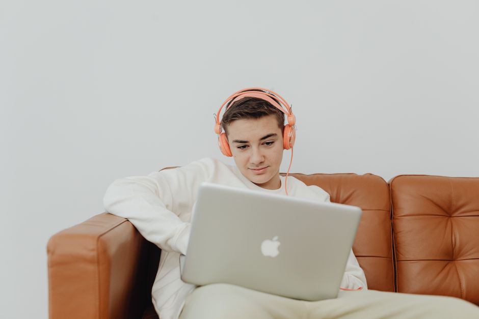 Jeune homme assis sur un canapé, utilisant un ordinateur portable avec un casque et écoutant de la musique à l'intérieur.
