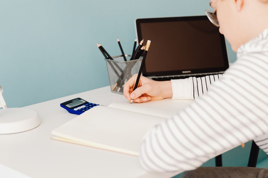 Jeune adulte écrivant avec des crayons à un bureau avec ordinateur portable et calculatrice, idéal pour les thèmes d'étude et de travail.
