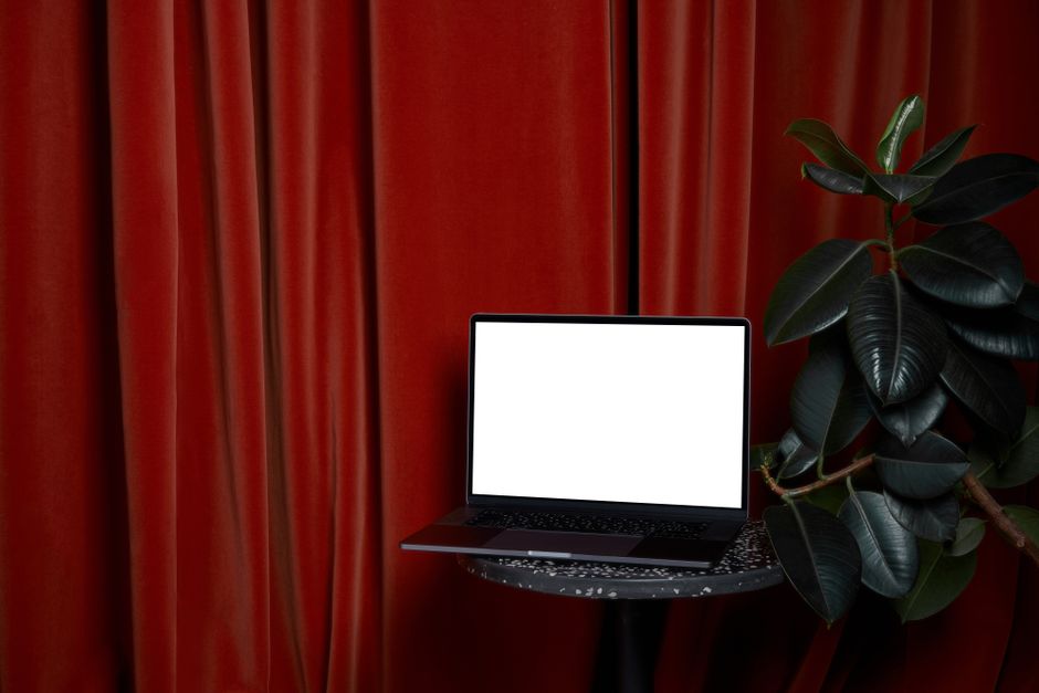 Ordinateur portable à écran blanc posé sur une table ronde près d'une plante aux feuilles vertes, avec des rideaux rouges en arrière-plan.