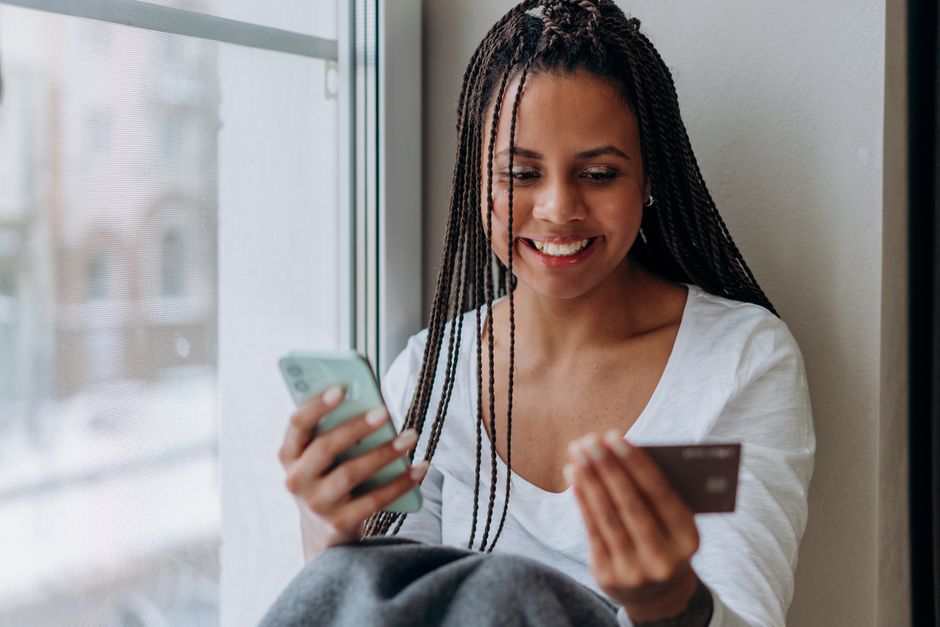 Femme souriante utilisant un smartphone et une carte de crédit pour faire des achats en ligne à l'intérieur, sous une lumière naturelle.