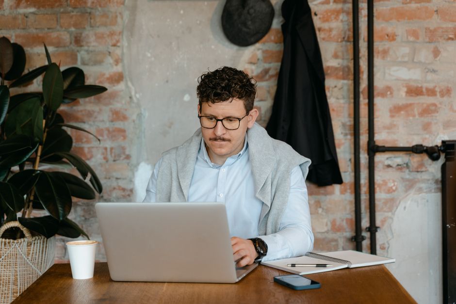 Homme à lunettes travaillant sur un ordinateur portable à une table en bois dans un bureau rustique.