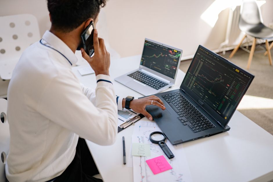 Homme tenant un téléphone, travaillant sur plusieurs ordinateurs portables avec des graphiques financiers à un bureau.