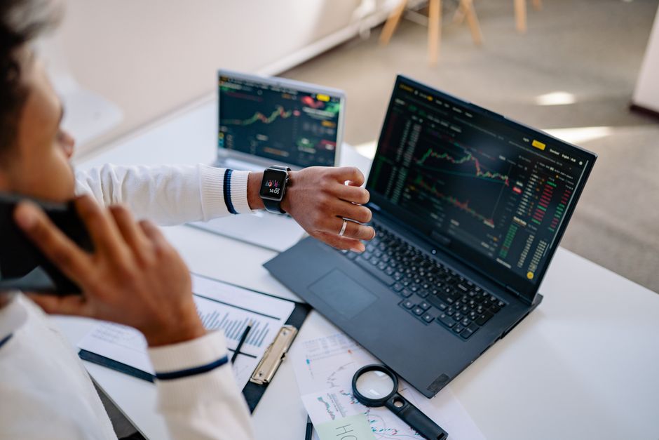Homme multitâche utilisant une montre connectée et un ordinateur portable pour analyser des graphiques boursiers au bureau.