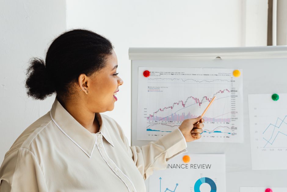 Femme pointant un graphique boursier sur un tableau blanc, expliquant des tendances financières.