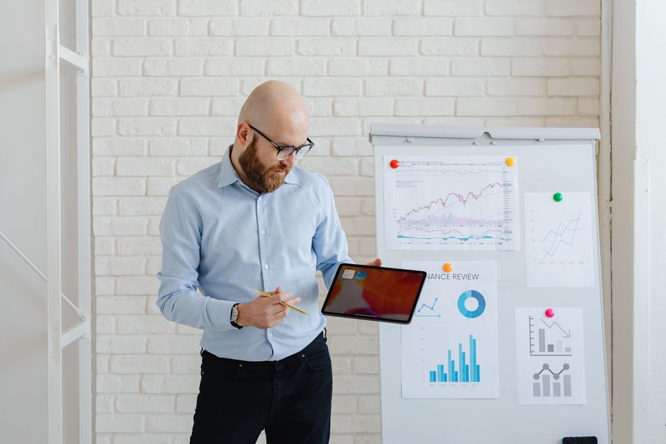 Un homme chauve présentant des données financières à l'aide d'une tablette et de graphiques sur un tableau blanc.