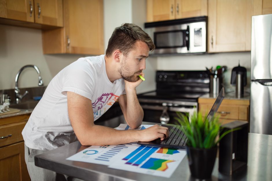 Jeune homme travaillant à distance sur un ordinateur portable dans une cuisine, concentré sur des graphiques de données.