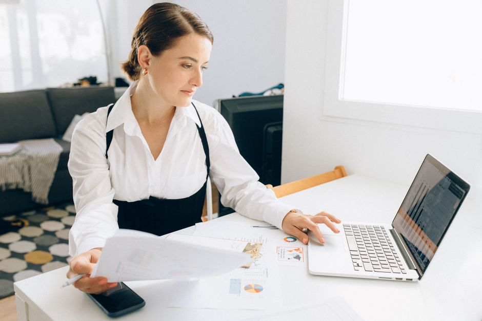 Femme à son bureau consultant des rapports d'activité sur un ordinateur portable, tenant des documents dans un environnement professionnel moderne.