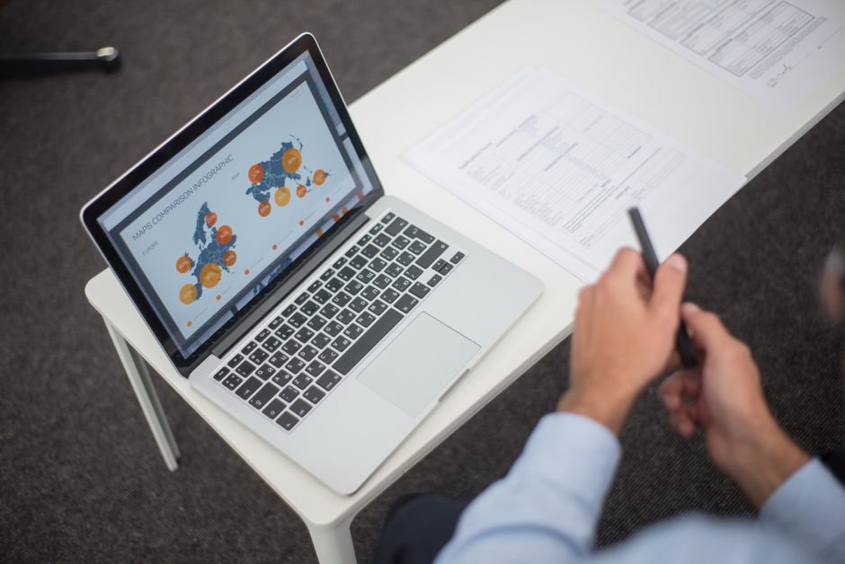 Professionnel analysant des infographies de données sur un ordinateur portable à un bureau, avec des documents et un stylo.